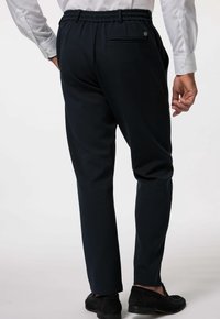 Mężczyzna w czarnych spodniach elastycznych z pasem i czarnych mokasynach, stojący z rękami swobodnie opuszczonymi wzdłuż ciała, widoczny z tyłu.