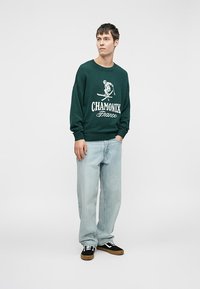 Zelena pletena jopica z belim grafičnim napisom "CHAMONIX Francija", sproščen kroj, v kombinaciji s svetlo modrimi kavbojkami in črnimi supergami z belimi detajli.