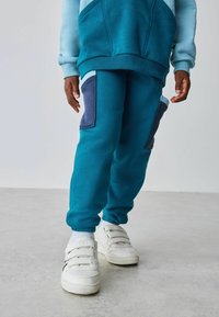 Sudadera verde azulado con detalles en azul claro, combinada con pantalones jogger verde azulado que tienen bolsillos en marino. Zapatillas blancas con correas de Velcro completan el atuendo.