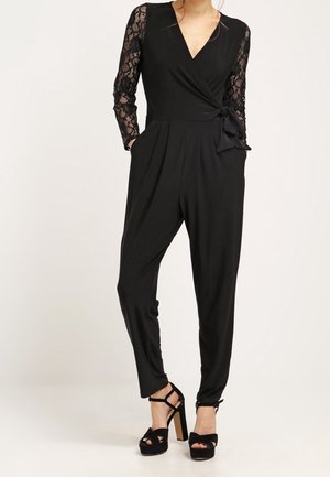 Kvinde iført en sort jumpsuit med blonder på de lange ærmer, slå-om-front, sidelommer, tilspidsede ben og sorte højhælede platform-sandaler.