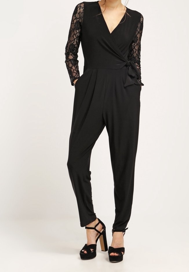 Femme portant une combi-pantalon noire avec manches longues en dentelle, devant croisé, poches latérales, jambes fuselées, et sandales noires à plateforme à talons hauts.