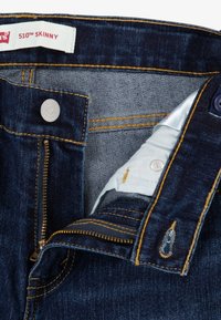 Jeans ajustados de color azul oscuro con costuras doradas visibles, cremallera plateada y botón de metal. El interior presenta una tela azul clara con una etiqueta.