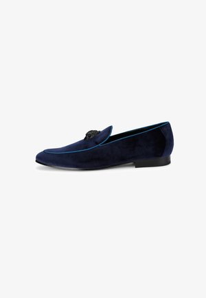 Marineblauwe suède loafer met zwarte zool en decoratief zwart kralenembleem op het bovenwerk, voorzien van blauwe bies langs de randen.