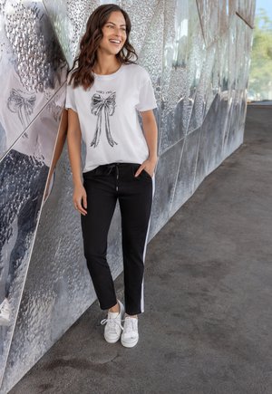 White t-shirt with black bow graphic, paired with black jogger pants featuring white side stripes. Worn with white sneakers, against a textured background.