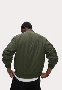 Giacca bomber verde oliva con maniche lunghe, polsini e orlo a coste, tessuto leggero e zip frontale.