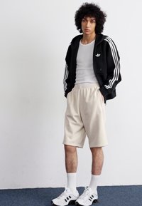 Sweatshirt preto com fecho, três riscas brancas, top branco, calções bege e ténis brancos com detalhes pretos; de pé sobre um tapete azul.