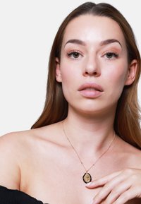 Collana in oro con ciondolo ovale caratterizzato da dettagli intricati e bordo in pietra nera. Il modello indossa un top a spalle scoperte.