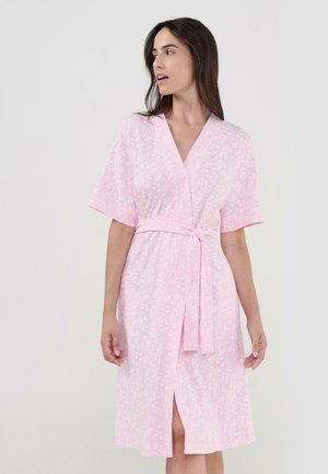 Mujer con una bata floral rosa claro con cinturón y mangas cortas, de pie y mirando hacia su izquierda contra un fondo sencillo.