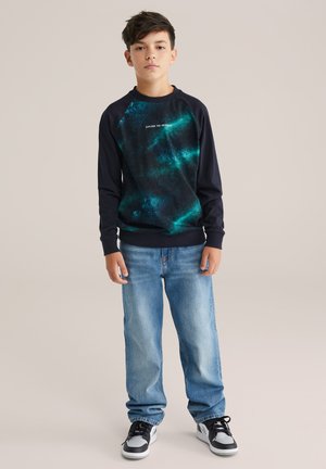 Dunkelblauer Sweatshirt mit einem grünen Galaxie-Print und weißem Text, kombiniert mit hellblauen Jeans und schwarzen Sneakers mit weißen Akzenten.