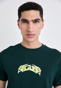 Dark green cotton T-shirt featuring a prominent yellow graphic design on the front, with a classic crew neck and short sleeves.