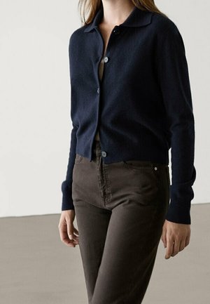 Femme portant un cardigan bleu marine boutonné et un pantalon taille haute marron foncé, debout devant un fond clair uni.
