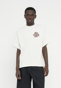 Λευκό υπερμεγέθες κοντομάνικο t-shirt με το γραφικό "Live On Air" σε καφέ χρώμα, συνδυασμένο με σκούρο παντελόνι. Λείο υφασμάτινο υφή.