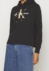 Svart hoodie med en framsidesgrafik i beige och vitt som säger "Calvin Klein Jeans" samt en liten logotyp på ärmen. Blandning av bomull.