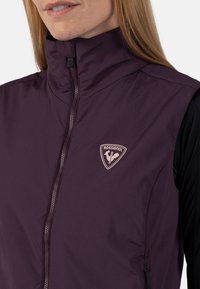 Ärmellose Damenweste aus dunkelviolettem Stoff, mit hohem Kragen, Frontreißverschluss und einem Logo-Patch in Hellrosa. Glatte Textur.
