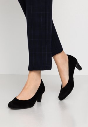 Escarpins en daim noir à talon bas portés avec un pantalon court à carreaux bleu foncé sur un sol gris clair.