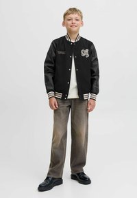 Giacca varsity nera con maniche in pelle, polsini a righe e un design ricamato. Abbinata a pantaloni larghi marroni sbiaditi e scarpe nere.