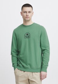 Felpa verde realizzata in tessuto morbido, con scollatura rotonda e un piccolo motivo stampato nero al centro. Maniche lunghe con polsini a coste.