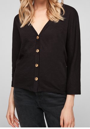 Žena v černém svetru s výstřihem do V na knoflíky s knoflíky ve vzoru želvoviny a černých džínách, stojící před jednobarevným pozadím.