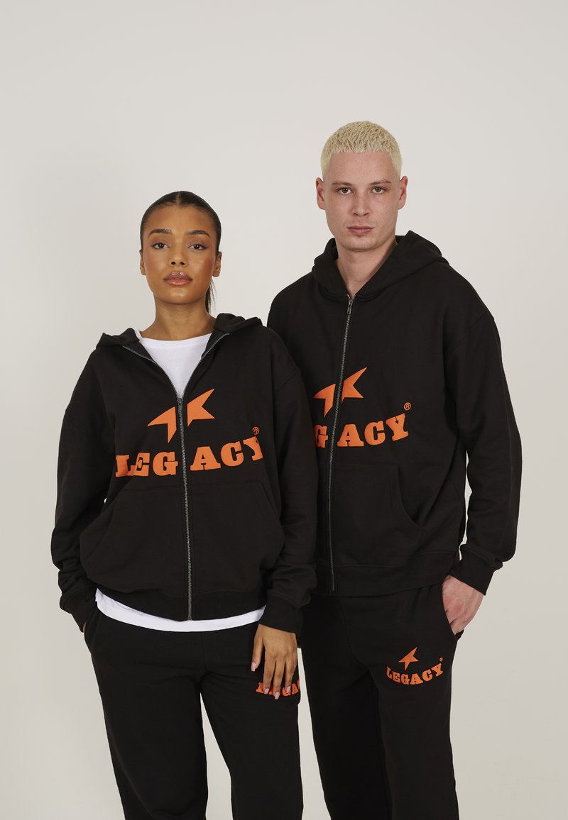 Black zip-up hoodies with orange "LEGACY" text and logo, paired with matching black sweatpants. Fabric appears smooth, relaxed fit.
