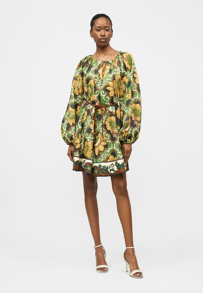 Γυναίκα που φοράει μακρυμάνικο φόρεμα με ζώνη, διακοσμημένο με πράσινα, κίτρινα και καφέ ανθισμένα μοτίβα, και λευκές ψηλοτάκουνες σανδάλες, στέκεται μπροστά σε απλό φόντο.