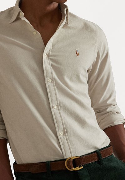 Chemise boutonnière beige clair en tissu texturé, arborant un petit logo brodé sur la poitrine. Assortie à une ceinture marron.