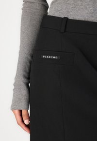 Svart skjørt med et elegant design, som har en frontlomme med en rektangulær etikett som står "BLANCHE" og en teksturert grå langermet topp.