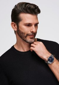 Orologio da polso da uomo con quadrante blu navy, cassa in acciaio inossidabile di colore argento e bracciale. Maglione a maglia nera con texture. Capelli corti e acconciati.
