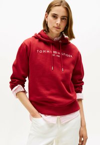 Rode katoenen hoodie met trektouwtjes, voorzien van witte tekst "TOMMY HILFIGER EST. 1985." Laagjes over een roze gestreept shirt, gecombineerd met witte broek.