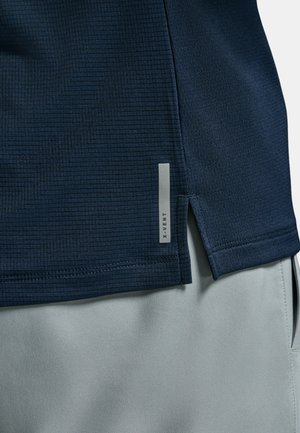 Detailní záběr na tmavě modrý strukturovaný lem košile s bočním rozparkem přes světle šedé kalhoty, s malou svislou reflexní značkou "X-VENT".