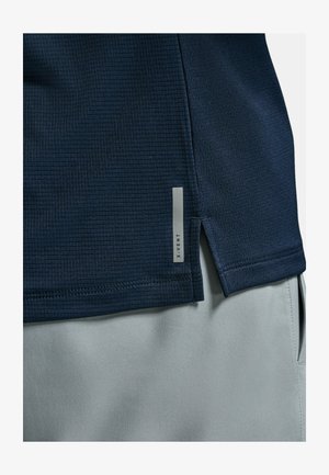 Detailní záběr na tmavě modrý strukturovaný lem košile s bočním rozparkem přes světle šedé kalhoty, s malou svislou reflexní značkou "X-VENT".