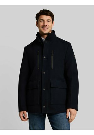 Smiling man wearing a black zip-up jacket with snap buttons and front pockets, standing against a plain gray background.