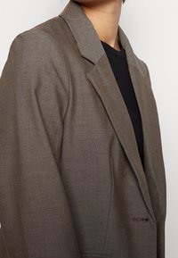 Ruskea räätälöity bleiseri, jossa on rakenteellinen kaulus ja sileä pinta, yksinkertainen nappisulku ja etutasku.