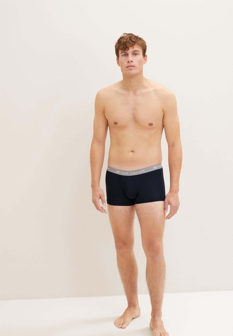 Čierne boxerky s šedým pásom, na ktorom je zobrazené meno značky, priliehavý strih a hladká textúra látky. Model stojí naboso.