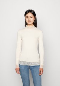 Langermet, ribbet topp i lys beige med blondedetaljer ved kragen og mansjettene, med en tettsittende design og myk tekstur.