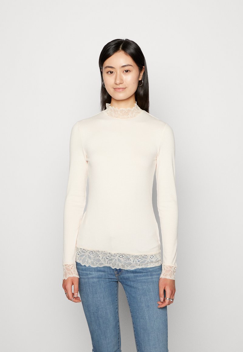 Langermet, ribbet topp i lys beige med blondedetaljer ved kragen og mansjettene, med en tettsittende design og myk tekstur.