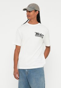 T-shirt bianco con stampa grafica nera e accenti a scacchi sul lato sinistro del petto. Indossato con jeans in denim blu, con una vestibilità rilassata.