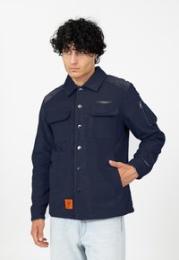 Marine jasje met knopen en twee voorzakken, met een oranje label. Materiaal heeft een gestructureerde afwerking en een ritszak op de mouw.