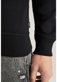 Sudadera negra con dobladillos y puños acanalados, con una pequeña etiqueta "JOOP!". Llevada sobre unos jeans gris texturizados con detalles desgastados.