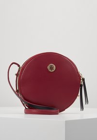 Borsa a tracolla rotonda in pelle rossa con hardware e cerniera dorati, caratterizzata da un emblema del marchio e una tracolla rimovibile. Superficie testurizzata.
