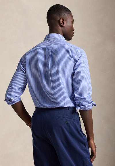 Άνδρας που φοράει μπλε και λευκό πουκάμισο με κατακόρυφες ρίγες, μανίκια γυρισμένα και σκούρο μπλε παντελόνι, στέκεται με την πλάτη στην κάμερα.