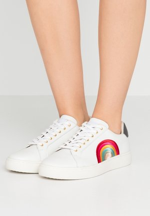 Zapatillas blancas con ojales dorados y bordado colorido de arcoíris, usadas sobre piernas desnudas contra un fondo gris liso.