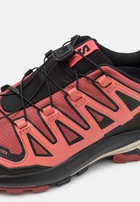 Chaussure de sport rouge et noire avec une tige texturée, un système de laçage élastique et une semelle Contagrip, dotée d'un panneau en maille respirante.