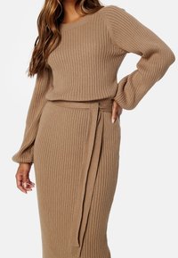 Femme portant un pull fin côtelé beige à manches longues et une jupe ceinturée assortie, posant avec une main sur la hanche et l'autre détendue.