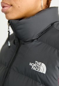Black puffer jacket with a high collar, featuring a front zipper and white embroidered logo on the chest. Smooth, insulated fabric.