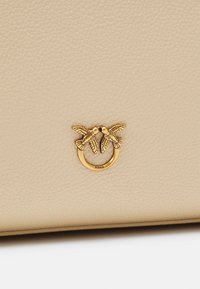 Béžová kožená kabelka s textúrovaným povrchom a detailom v zlatej farbe, ktorý zobrazuje dvoch vtákov v kruhovej obrube.