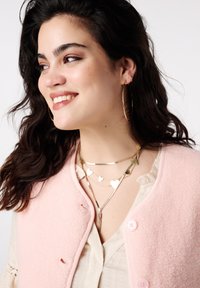 Roze fleecevest met ronde knopen, gedragen over een lichtbeige shirt. Gouden kettingen met hartvormige ontwerpen en grote oorringen.