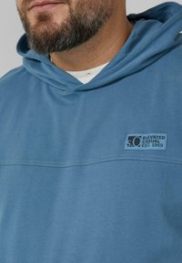 Niebieska bluza z miękkiego materiału, z naszywką z logo na przodzie, przeszyciami na klatce piersiowej oraz kapturem ze sznurkiem.