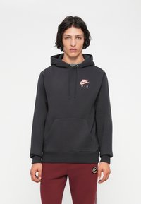 Jovem vestido com um hoodie preto da Nike e calças burgundy com o logótipo da Nike, em pé contra um fundo branco simples.