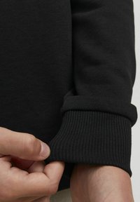 Felpa nera con polsini a coste, texture liscia e una vista ravvicinata di una mano che regola le maniche.