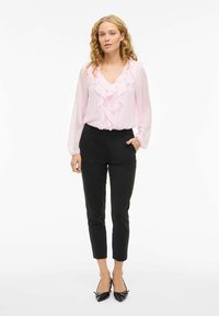 Gaiši rozā blūze ar frillēm priekšpusē, garām piedurknēm un melniem piegrieztiem biksēm. Melni kurpes ar asu purngalu noslēdz apģērbu.